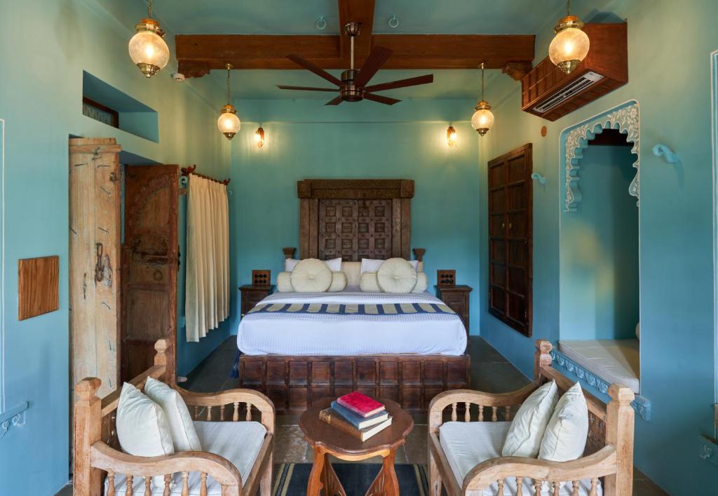 9 Iconic Heritage Hotels in Udaipur 22 A cozy bedroom in the style of heritage hotels in Udaipur, with carved wooden furniture, a large bed, blue walls, hanging pendant lights, and a ceiling fan. A small table with books sits between two elegant chairs.