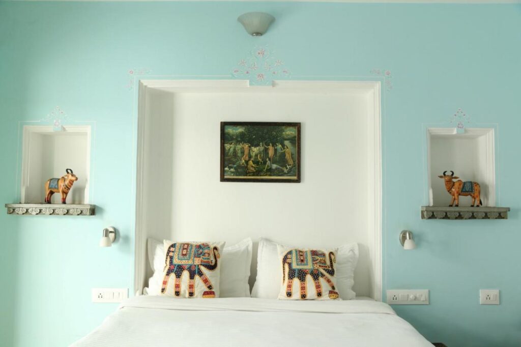 9 Iconic Heritage Hotels in Udaipur 3 A neatly made bed with white linens and two elephant-patterned pillows stands against a light blue wall, echoing the charm of heritage hotels in Udaipur. Decorative cow figurines line the shelves, with a framed painting above the bed.