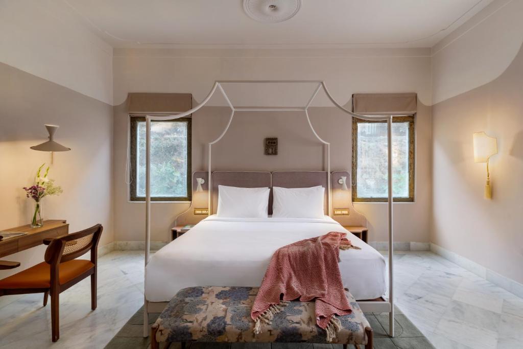 9 Iconic Heritage Hotels in Udaipur 7 A bright, elegant bedroom in one of the charming heritage hotels in Udaipur, featuring a four-poster double bed with white linens and a pink throw, two windows, a wooden desk with chair, wall lamp, and a patterned bench at the foot of the bed.