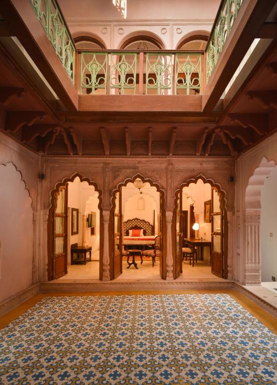 An ornate interior courtyard reminiscent of boutique hotels in New Delhi, with patterned blue and yellow floor tiles, arched doorways, wooden accents, and a green-railed balcony above. The warm space features inviting traditional decor.