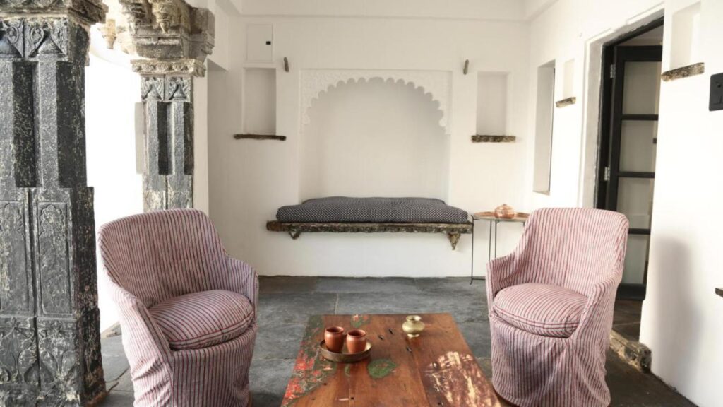 9 Iconic Heritage Hotels in Udaipur 6 A cozy sitting area inspired by heritage hotels in Udaipur, with striped armchairs, a rustic wooden table with copper tea set, a cushioned bench, and ornate stone columns set against white walls in a bright, minimalist room.