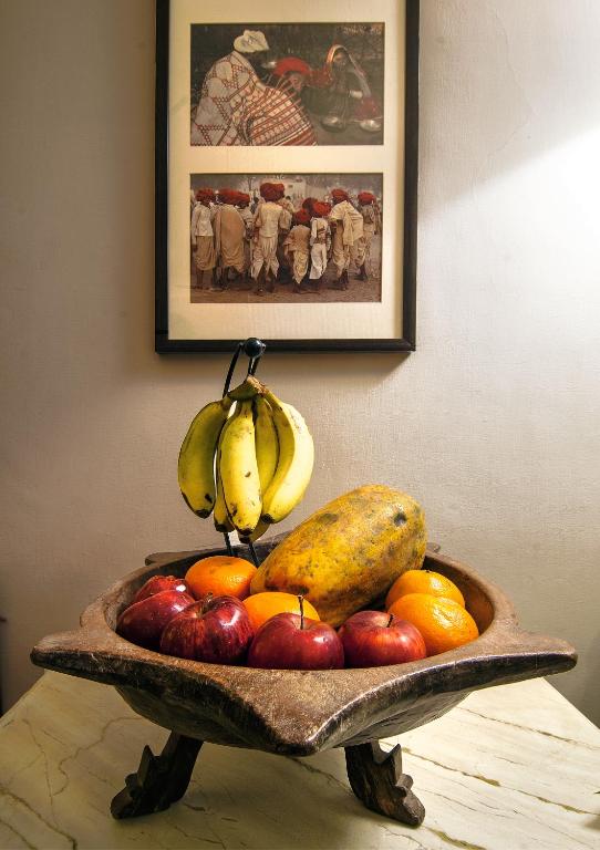 A wooden fruit bowl on a table holds bananas, a papaya, apples, and oranges—a scene often found in boutique hotels in New Delhi. Behind it, two framed pictures of people in traditional clothing hang on a cream-colored wall.