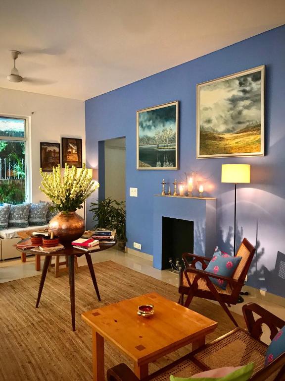 A cozy living room with a blue accent wall, fireplace, artwork, and wooden furniture evokes the charm of boutique hotels in New Delhi. Sunlight streams in from a large window, highlighting a round table with yellow flowers, candles, and a floor lamp.
