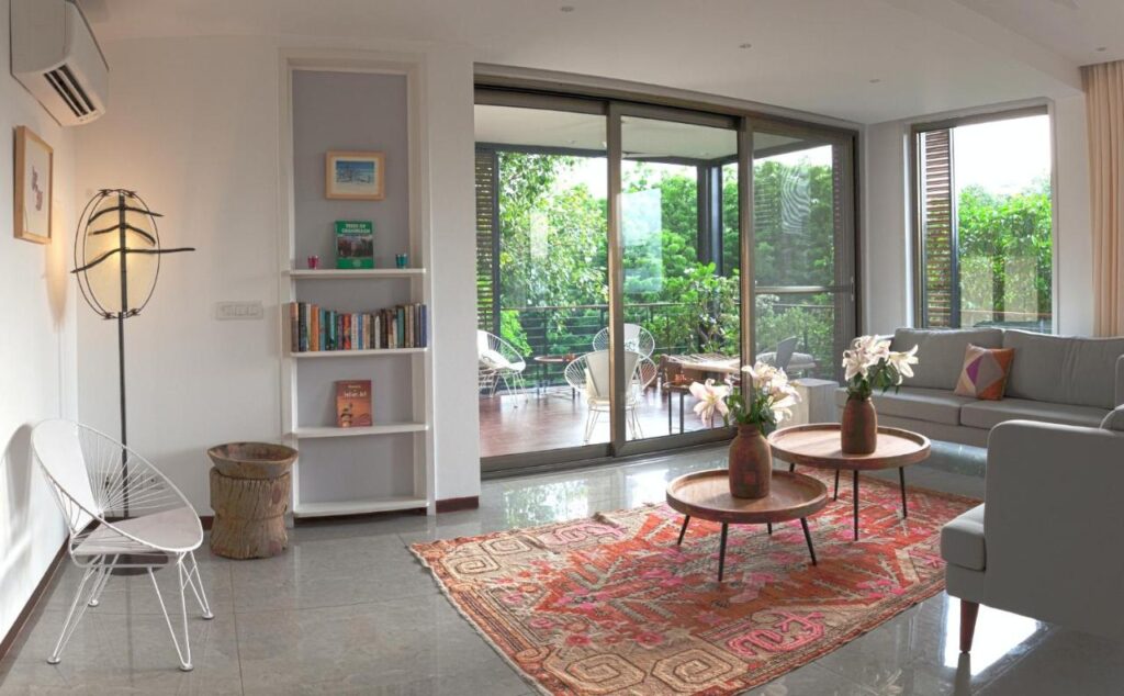 Bright, modern living room with large glass doors opening to a green patio. Reminiscent of boutique hotels in New Delhi, there’s a gray sofa, round coffee tables with flowers, a white chair, mixed decor, bookshelves, and a patterned rug on tiled flooring.