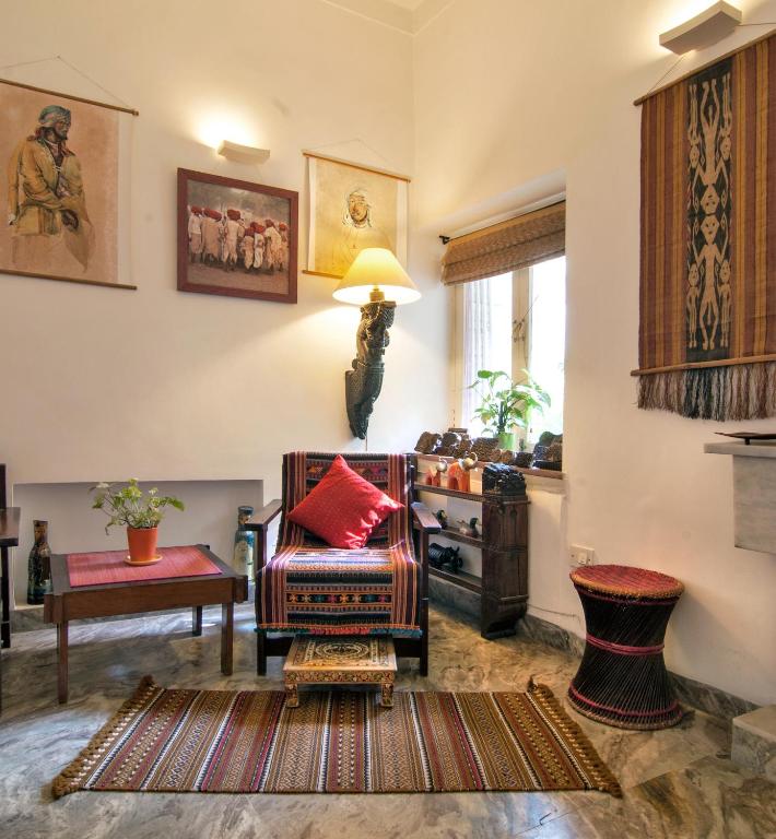 A cozy living room with ethnic decor, reminiscent of boutique hotels in New Delhi, featuring a patterned armchair and footstool, a small table with a potted plant, wall art, woven wall hanging, window plants, and warm lighting.