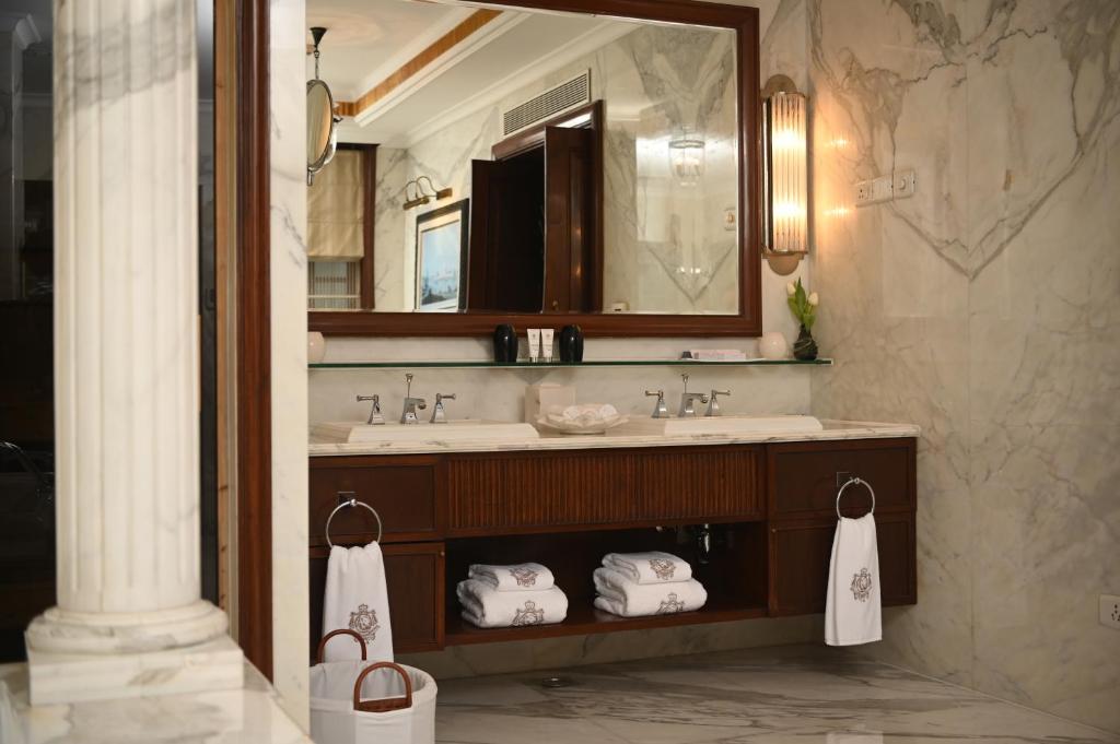 Luxurious marble bathroom with double sinks, a large mirror, wall sconces, rolled white towels, and wooden cabinets—reminiscent of boutique hotels in New Delhi. A decorative column and toiletries add to the elegant, upscale atmosphere.