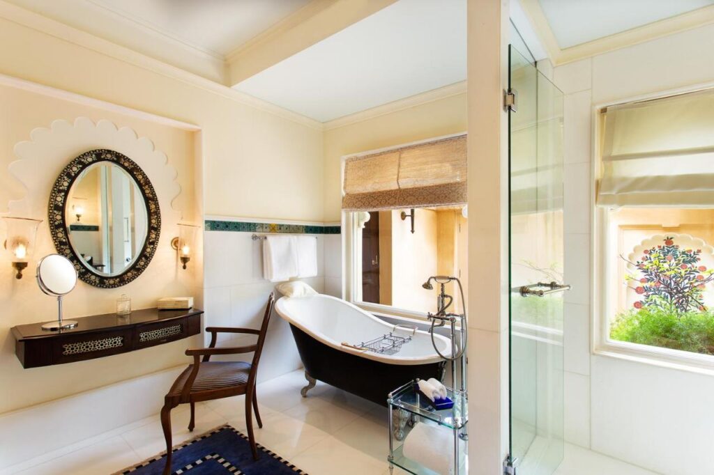 9 Iconic Heritage Hotels in Udaipur 26 Elegant bathroom with a black clawfoot bathtub, glass shower, large window with a blind, wooden chair, and wall-mounted sink under a round mirror—evoking the luxurious style of heritage hotels in Udaipur, softly illuminated by natural light.