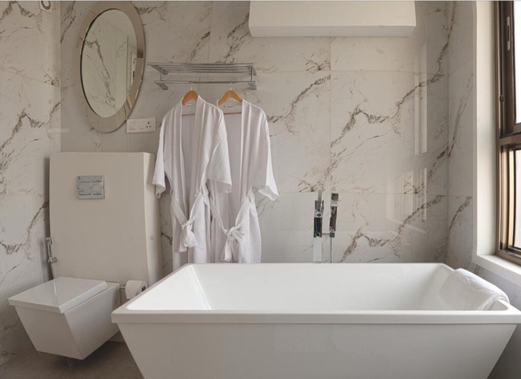 Modern bathroom with marble walls, a rectangular bathtub, wall-mounted toilet, two white bathrobes on hangers, a round mirror, and a large window—evoking the elegance found in boutique hotels in New Delhi.