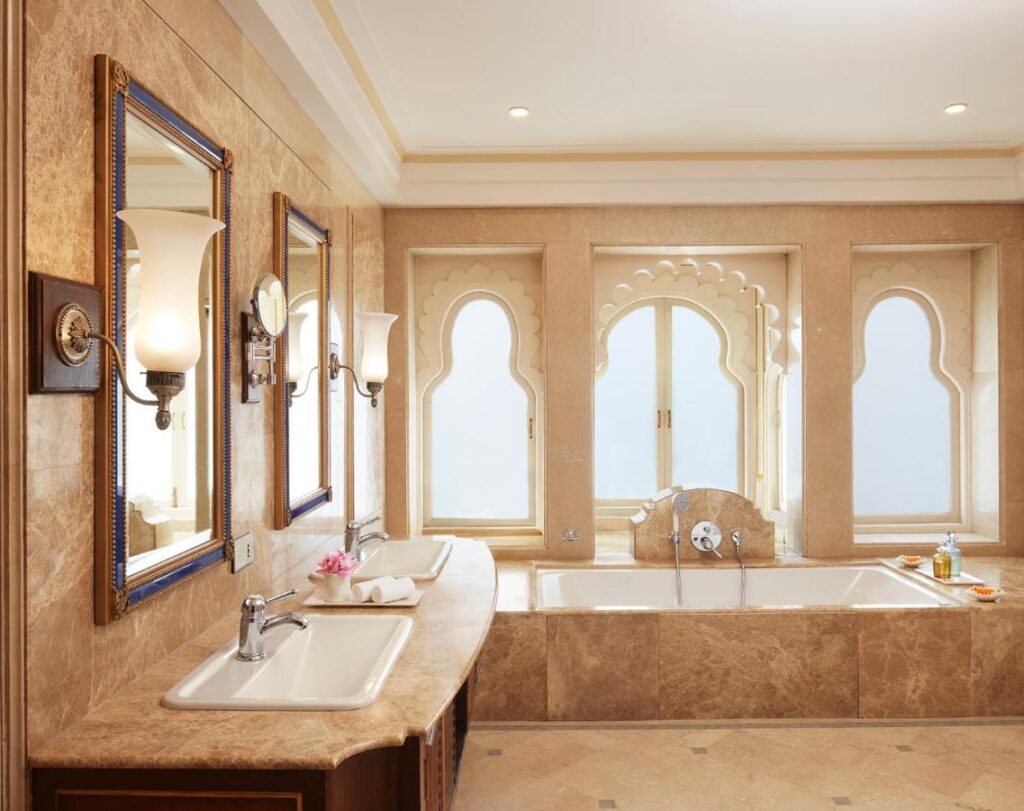 9 Iconic Heritage Hotels in Udaipur 14 A luxurious bathroom inspired by heritage hotels in Udaipur, featuring a marble double sink, large mirrors, elegant sconces, a spacious bathtub, and arched windows with intricate detailing and frosted glass.