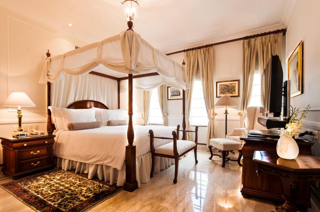 Luxurious bedroom inspired by boutique hotels in New Delhi, featuring a four-poster bed, classic wooden furniture, elegant lamps, a patterned rug, and large windows with cream curtains letting in natural light.