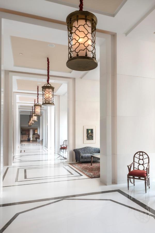 A bright, elegant hallway inspired by boutique hotels in New Delhi, with white walls and floor, hanging lantern-style lights, a patterned rug, a dark sofa, framed artwork, and a decorative wooden chair.
