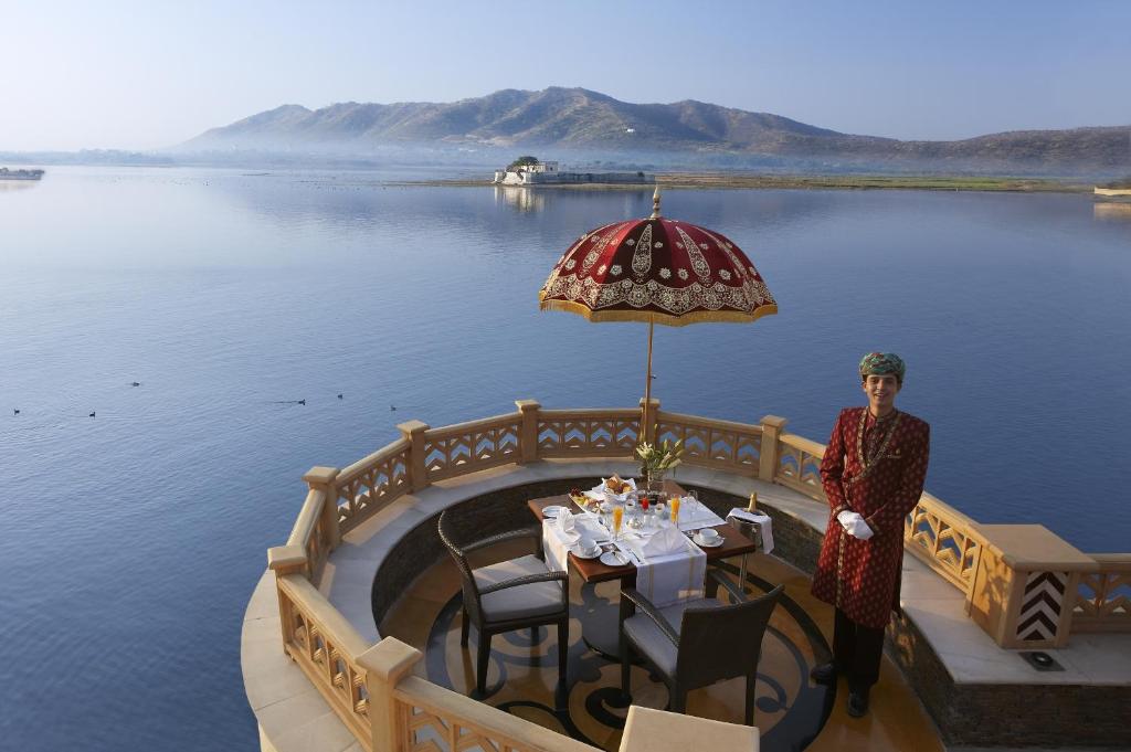 9 Iconic Heritage Hotels in Udaipur 30 A traditionally dressed server stands beside a breakfast table set for two on a balcony at one of the heritage hotels in Udaipur, overlooking a calm lake with distant hills, under a decorative red umbrella.