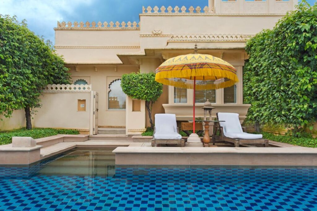 9 Iconic Heritage Hotels in Udaipur 23 A serene courtyard at one of the heritage hotels in Udaipur features a small blue-tiled pool, two white lounge chairs under a yellow umbrella, manicured trees, and an ornate cream-colored building in the background.