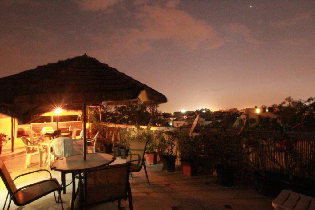 A rooftop terrace at dusk in one of the boutique hotels in New Delhi features tables and chairs under a thatched umbrella, potted plants, and soft lighting. The sky glows purple and orange as distant city lights twinkle on the horizon.