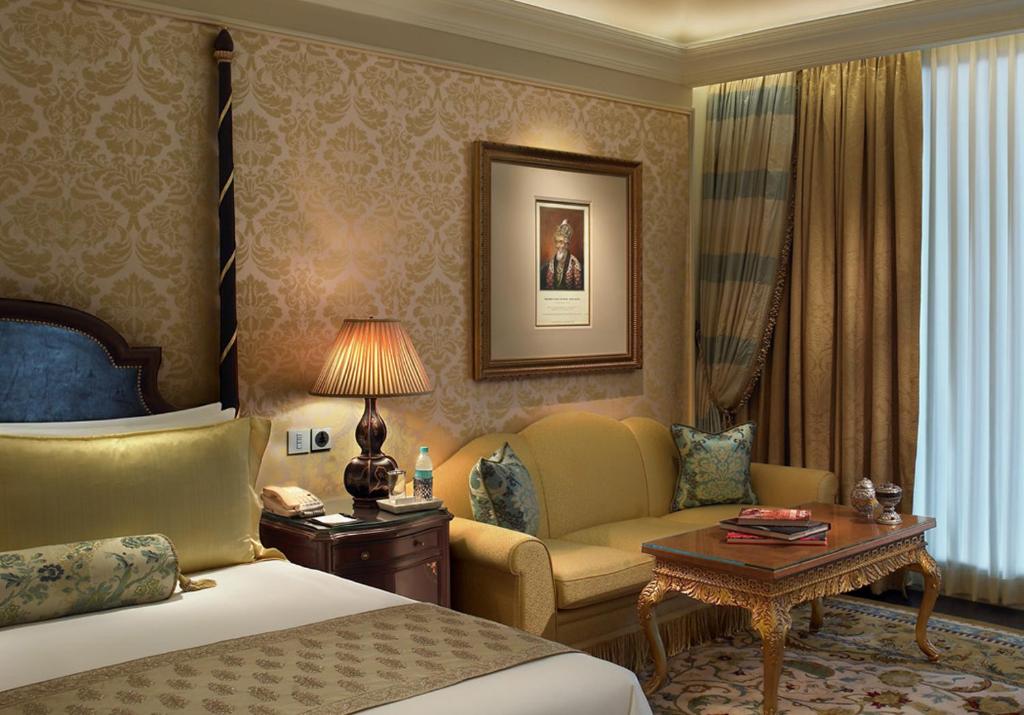 Luxurious boutique hotel room in New Delhi with ornate wallpaper, a bed with decorative pillows, a yellow sofa, an elegant wooden coffee table, and a table lamp. A framed picture and draped curtains add to the sophisticated decor.