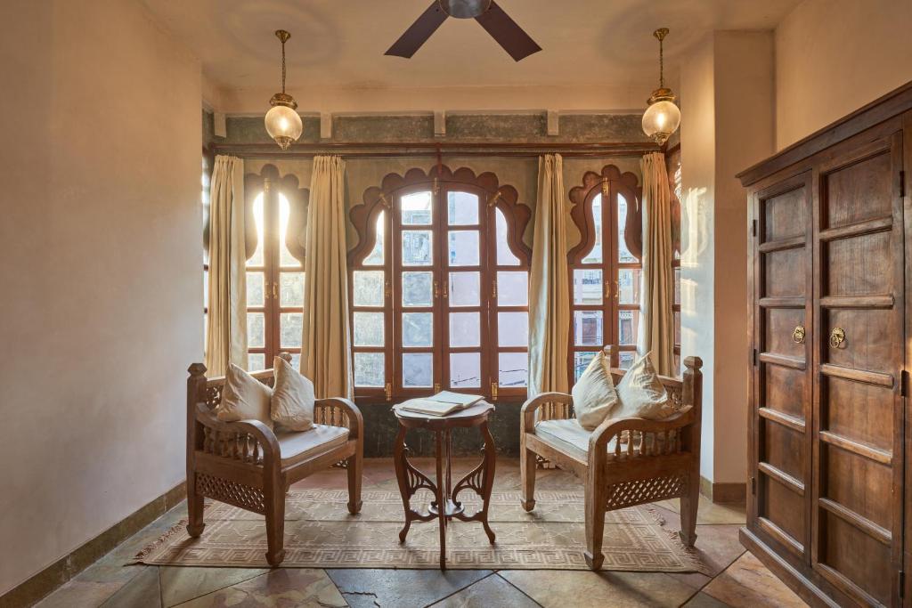 9 Iconic Heritage Hotels in Udaipur 19 A cozy sitting area, reminiscent of heritage hotels in Udaipur, features two wooden armchairs, a small round table, and large arched windows with sheer curtains letting in sunlight. A ceiling fan and wooden wardrobe complete the elegant room.