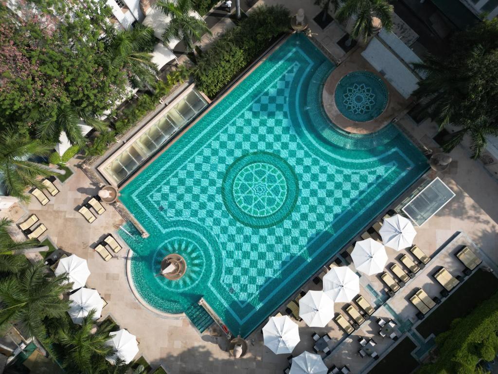 Aerial view of an ornate, turquoise swimming pool with geometric patterns at one of the boutique hotels in New Delhi, surrounded by lounge chairs, white umbrellas, palm trees, and lush greenery in a luxurious outdoor setting.
