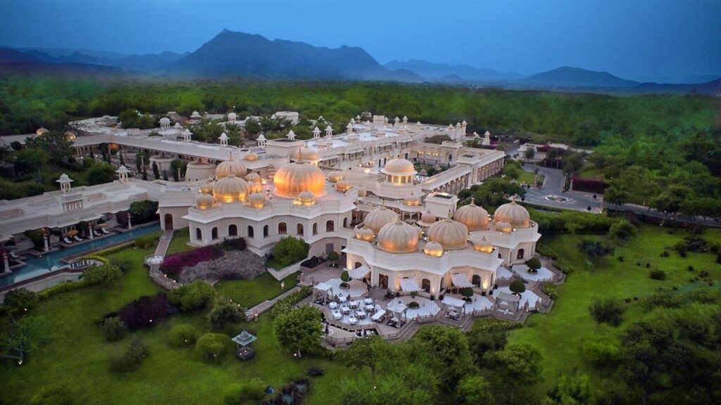 9 Iconic Heritage Hotels in Udaipur 25 Aerial view of a grand palace-style hotel, reminiscent of heritage hotels in Udaipur, with multiple domes and ornate architecture, surrounded by lush greenery and gardens, set against distant mountains at dusk.
