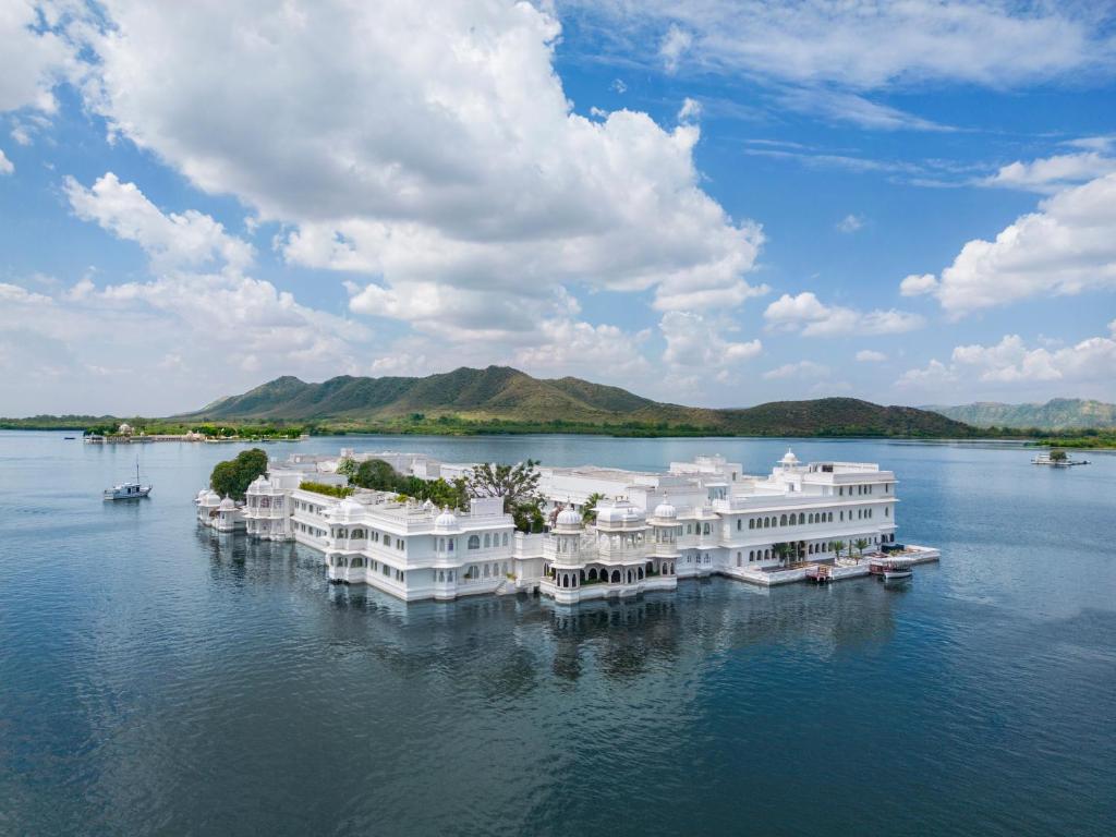 9 Iconic Heritage Hotels in Udaipur 32 A large white palace, one of the renowned heritage hotels in Udaipur, sits on the calm waters of a lake, surrounded by mountains and a partly cloudy blue sky, with small boats nearby.