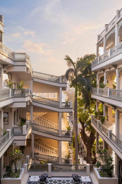 9 Iconic Heritage Hotels in Udaipur 9 A multi-story building with open corridors, white railings, and a central courtyard filled with greenery and a tall palm tree—reminiscent of heritage hotels in Udaipur—bathed in soft sunlight under a pastel sky.