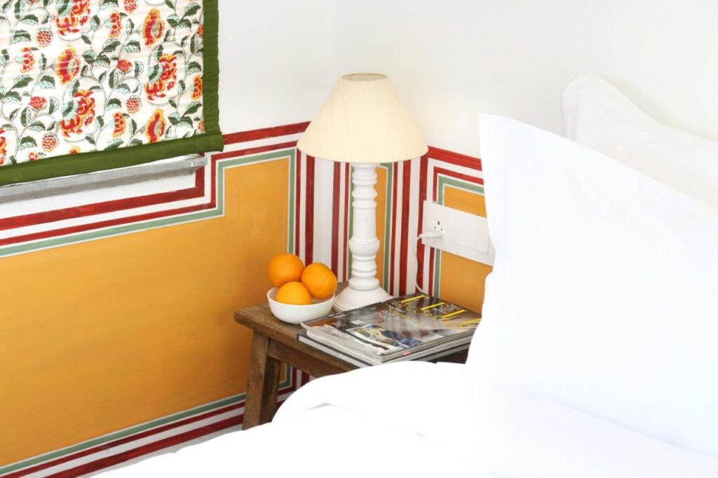 9 Iconic Heritage Hotels in Udaipur 5 A cozy bedroom corner reminiscent of heritage hotels in Udaipur, with a wooden side table holding a white lamp, a bowl of oranges, and magazines. The wall has colorful stripes, floral curtain above the table, and white bedding in the foreground.