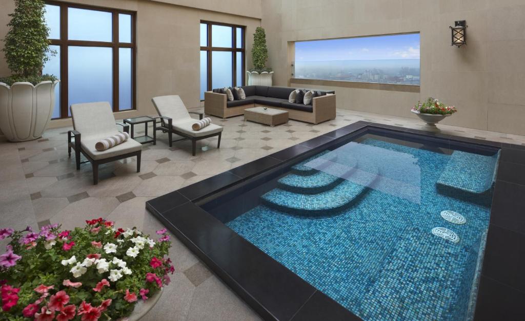 Luxurious indoor pool area in one of the finest boutique hotels in New Delhi, featuring a mosaic-tiled plunge pool, lounge chairs with rolled towels, a corner sofa, potted plants, fresh flowers, and large windows with city views.