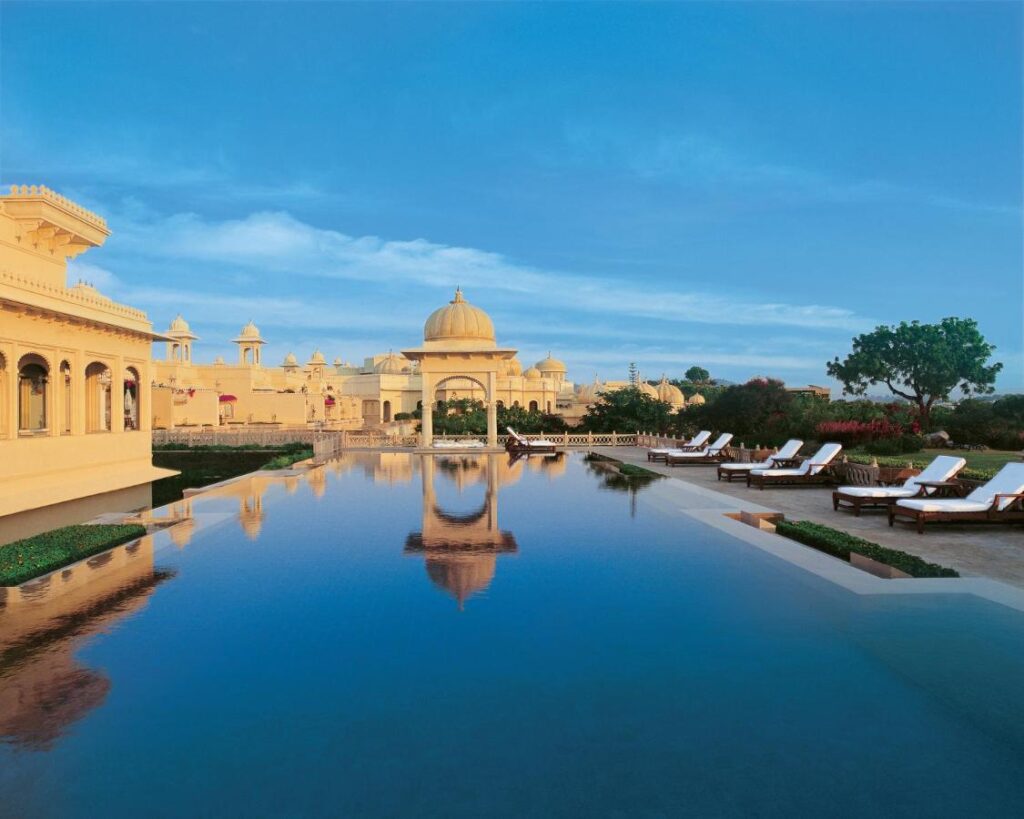 9 Iconic Heritage Hotels in Udaipur 24 A luxurious palace-style hotel, reminiscent of heritage hotels in Udaipur, with a large serene pool reflecting domed architecture, surrounded by lounge chairs and lush gardens under a clear blue sky.