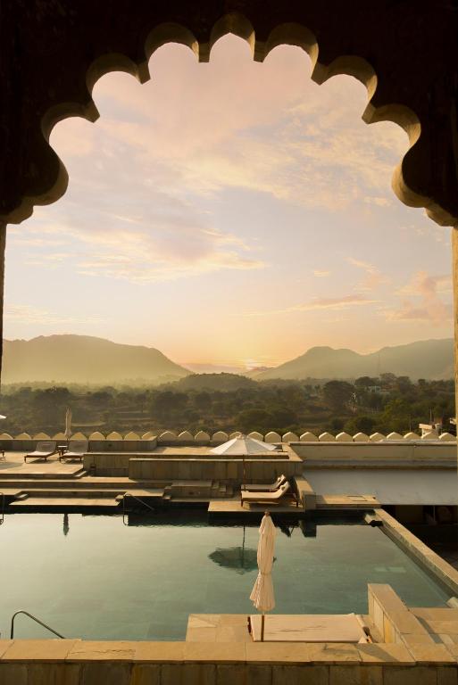 9 Iconic Heritage Hotels in Udaipur 36 A tranquil pool with sun loungers overlooks mountains and a valley, framed by an ornate arch at sunset, creating a serene and luxurious atmosphere reminiscent of heritage hotels in Udaipur.