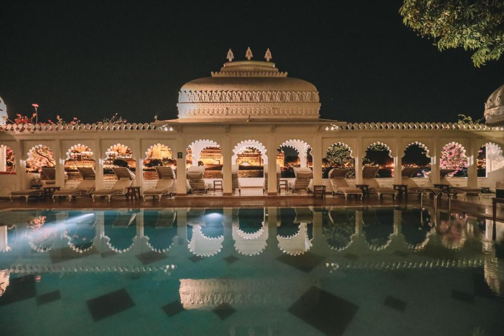 9 Iconic Heritage Hotels in Udaipur 33 Night view of an ornate heritage hotel in Udaipur with arched windows and intricate carvings, reflected in a calm swimming pool. Warm lights illuminate the structure, creating a serene and elegant atmosphere.