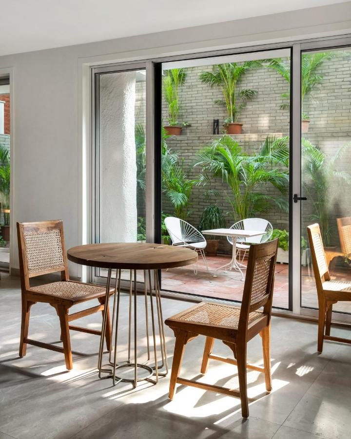 A round wooden table with three cane chairs set indoors near large glass doors, opening to a patio with white chairs and lush potted plants—reminiscent of the stylish interiors found in boutique hotels in New Delhi.