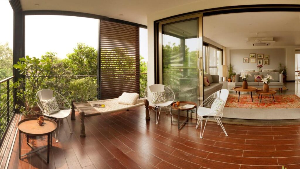 A spacious balcony with wooden flooring, white chairs, a charpoy, and small tables overlooks lush greenery—much like the serene escapes offered by boutique hotels in New Delhi—connected to a modern living room with large glass sliding doors, an orange rug, and wall art.