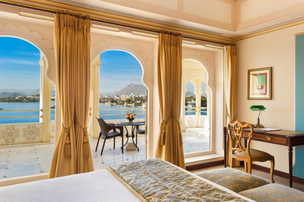 9 Iconic Heritage Hotels in Udaipur 13 A luxurious room in one of the heritage hotels in Udaipur, with gold curtains, a plush bed, wooden desk, and arched windows opening to a marble balcony overlooking a serene lake and distant mountains under a blue sky.