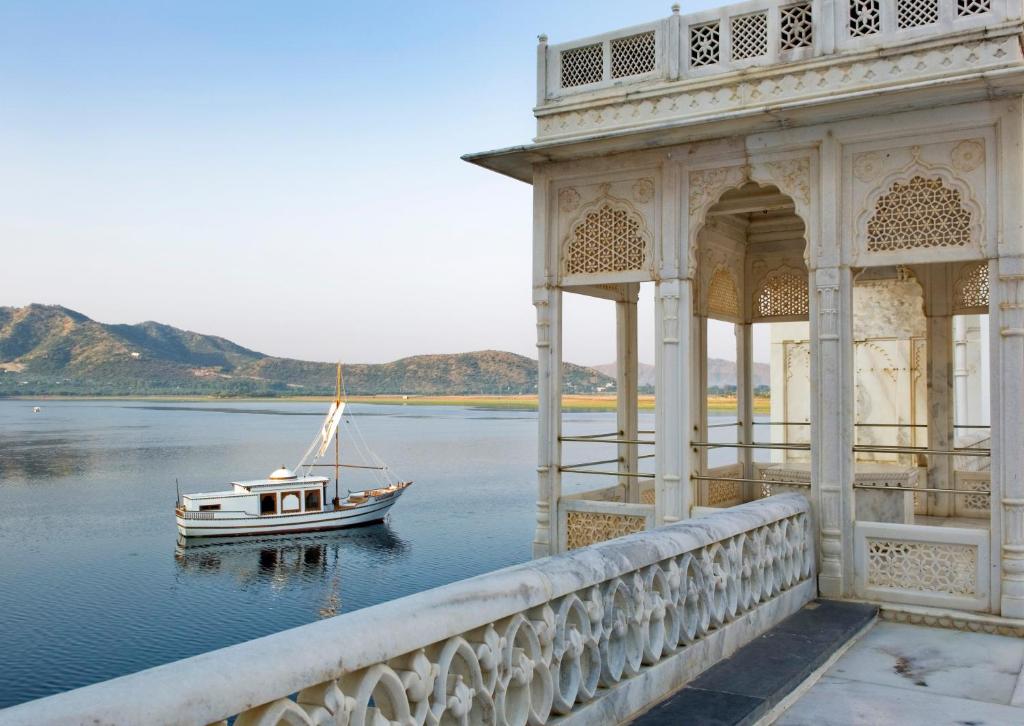 9 Iconic Heritage Hotels in Udaipur 31 A white marble pavilion with ornate carvings, like those at heritage hotels in Udaipur, overlooks a calm lake where a small sailboat floats near the shore. Distant hills rise under a clear sky in the background.