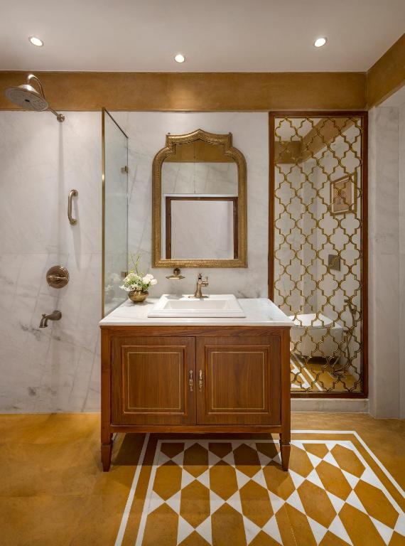 A bathroom inspired by boutique hotels in New Delhi, featuring a wooden vanity, marble countertop, ornate gold mirror, geometric gold-accented shower screen, and patterned tile floor. A vase with flowers sits on the counter.