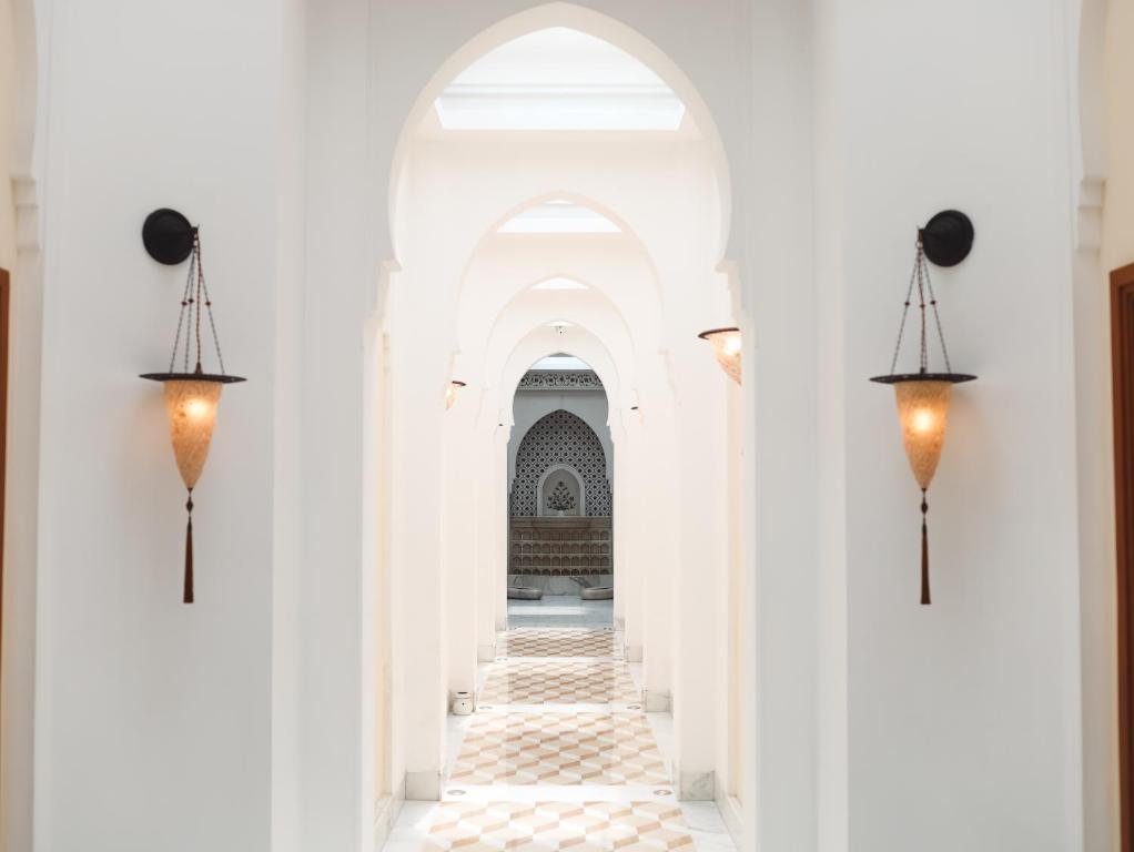 A bright corridor in one of the boutique hotels in New Delhi features white arched doorways, a patterned tile floor, and warm wall sconces leading to an ornate alcove with intricate designs.