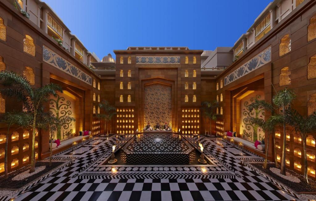 9 Iconic Heritage Hotels in Udaipur 29 A luxurious courtyard at heritage hotels in Udaipur boasts checkered black-and-white flooring, ornate golden walls, palm trees, and a central pool with decorative lanterns, all under a clear blue sky.