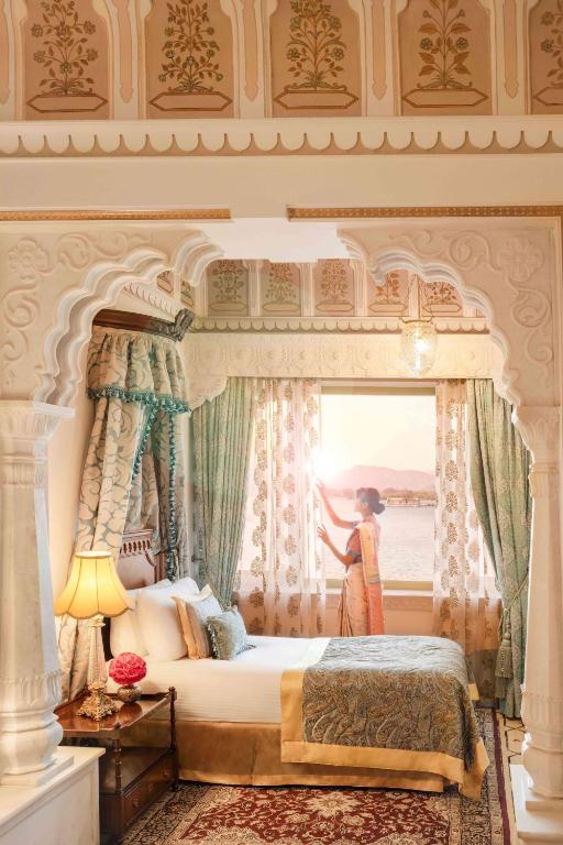 9 Iconic Heritage Hotels in Udaipur 34 A woman in traditional attire pulls back curtains in an ornately decorated room of a heritage hotel in Udaipur, with carved arches, a canopy bed, and patterned walls as sunlight streams through a window overlooking water and distant mountains.