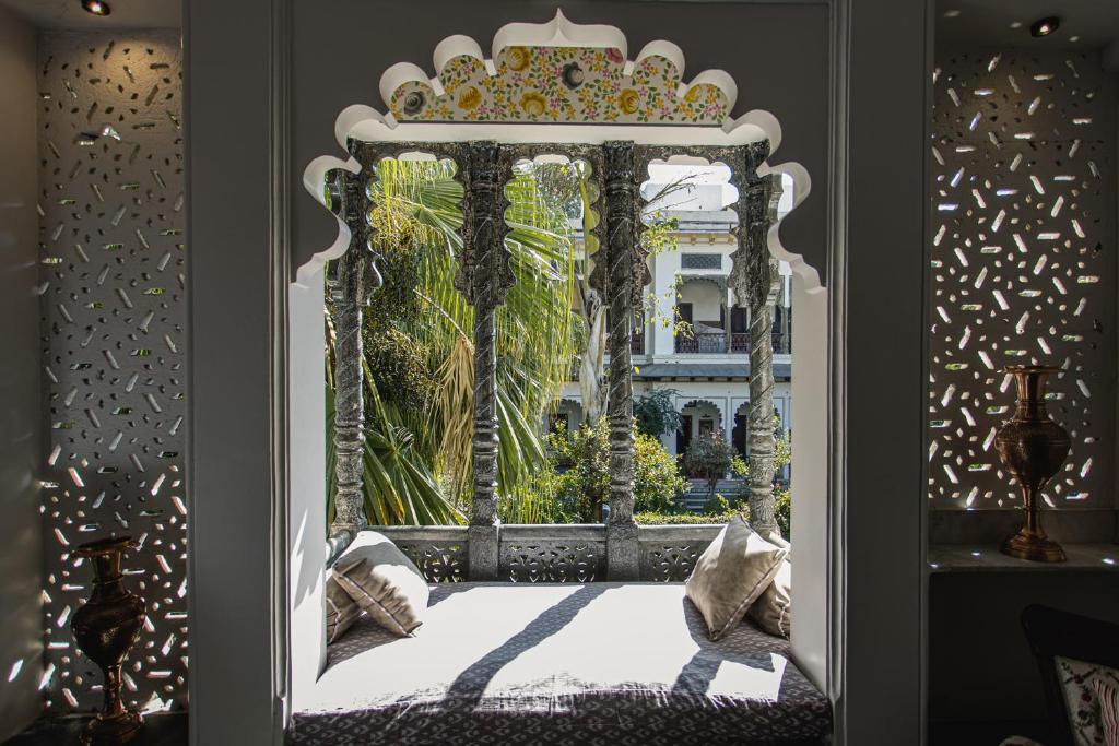 9 Iconic Heritage Hotels in Udaipur 18 Daybed with patterned cushions set in an ornate stone window alcove at one of the heritage hotels in Udaipur, overlooking a lush garden with palm leaves and traditional architecture. Sunlight casts intricate shadows on the walls.
