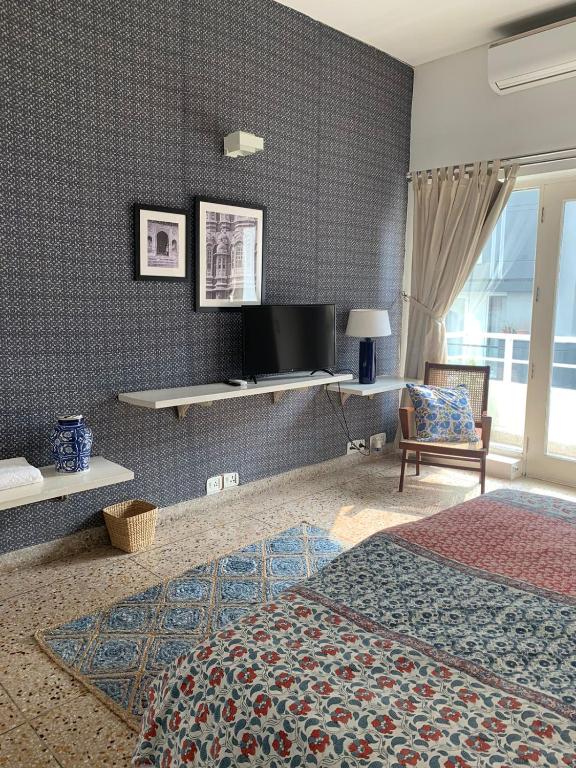 A cozy bedroom reminiscent of boutique hotels in New Delhi, featuring patterned wallpaper, framed pictures, a wall-mounted TV, a chair with colorful cushions, patterned bedding, a woven basket, a rug, and large windows letting in natural light.