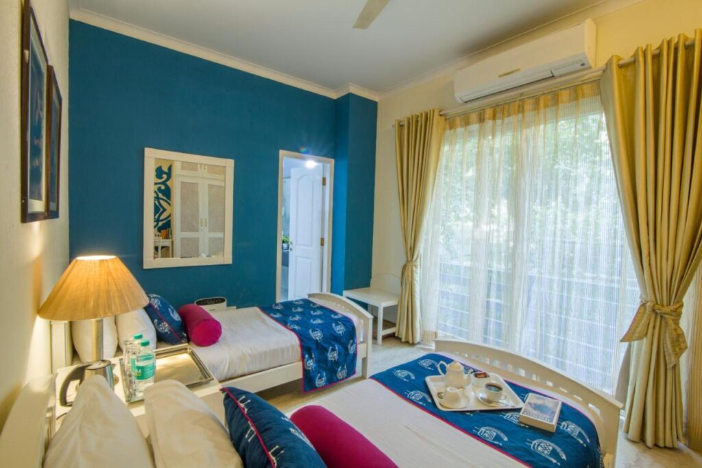 A bright, cozy bedroom with two single beds, blue accent wall, white furniture, lamp, large window with sheer curtains, air conditioner, and a tray with cups and a book—perfect for guests seeking boutique hotels in New Delhi.