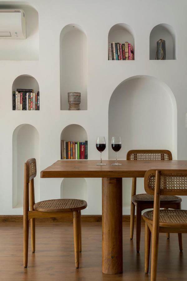 A modern dining area with a wooden table and three chairs, two glasses of red wine on the table, reminiscent of the chic interiors found in boutique hotels in New Delhi; white wall shelves hold books and decorative items.