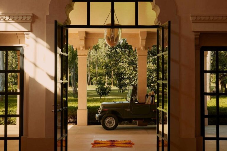 An open glass doorway leads to a veranda with a green jeep parked outside, surrounded by lush greenery and trees—offering the tranquil charm often found at boutique hotels in Jaipur. Sunlight casts warm shadows on the beige walls and tiled floor inside.