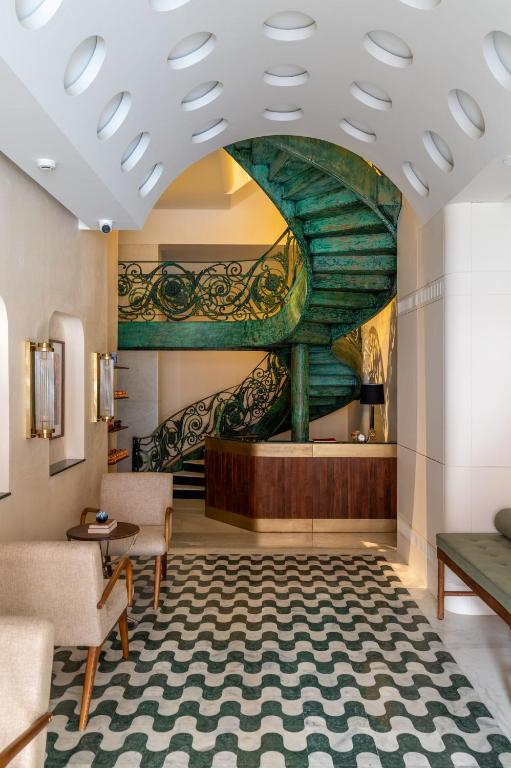12 BEST Boutique Hotels in Jaipur 23 A stylish lobby, reminiscent of boutique hotels in Jaipur, features a bold green spiral staircase with ornate railings, a wooden reception desk, modern armchairs, a green cushioned bench, and a wavy patterned floor in white and dark green tones.