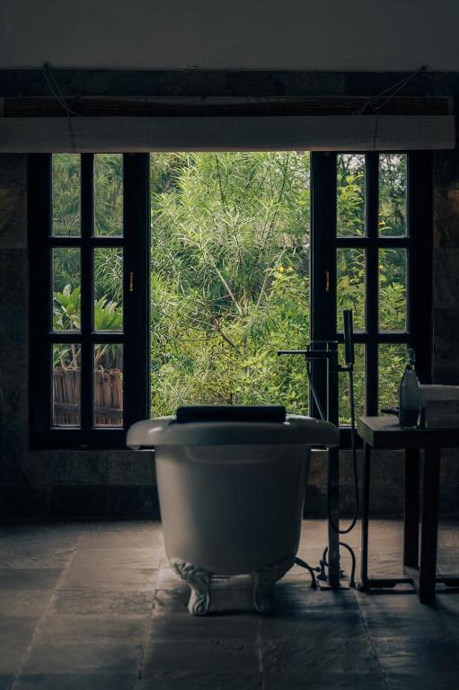 12 BEST Boutique Hotels in Jaipur 31 A freestanding bathtub sits in front of tall windows, overlooking lush green plants and trees outside—evoking the tranquil, inviting ambiance often found in boutique hotels in Jaipur. Soft natural light filters into the serene, dimly lit bathroom.