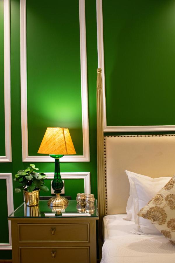 12 BEST Boutique Hotels in Jaipur 8 A bedside table in the style of boutique hotels in Jaipur, with a green lamp, a small potted plant, decorative jars, and a gold cup beside a cream-colored bed with white pillows against a bright green wall with white trim.
