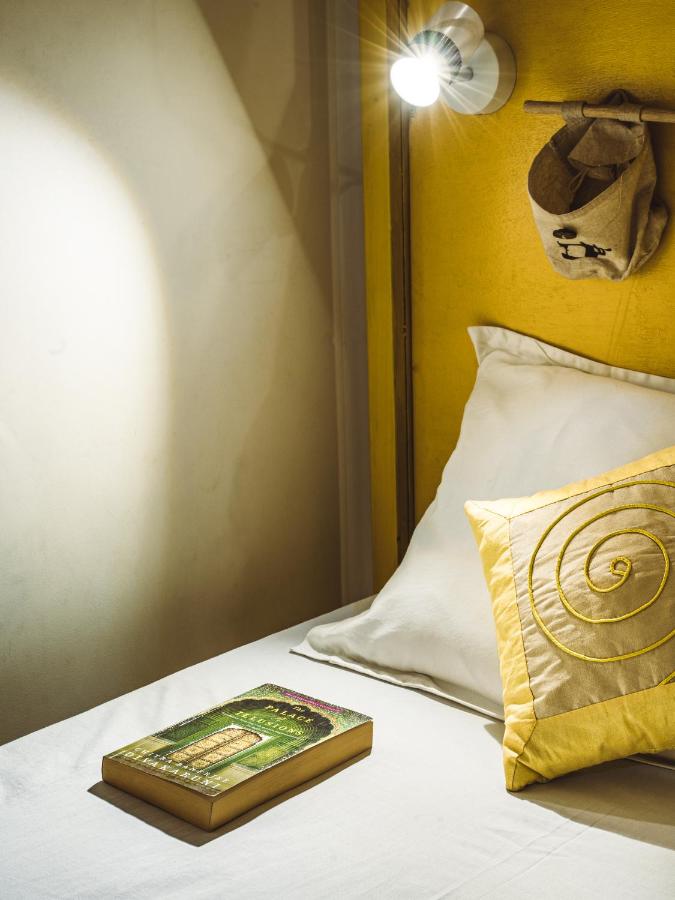 12 BEST Boutique Hotels in Jaipur 40 A cozy bed with white sheets, a yellow spiral cushion, and a book resting on the mattress—like those found in boutique hotels in Jaipur. A wall-mounted reading light shines above, and a small fabric pouch hangs on the yellow headboard.