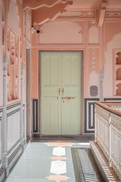 12 BEST Boutique Hotels in Jaipur 49 A pastel-hued hallway reminiscent of boutique hotels in Jaipur features ornate, decorative walls and a cream-colored double door at the end. Intricate latticework lines the right side, while soft sunlight casts patterns on the polished floor.