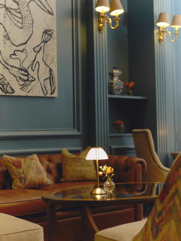 9 Unique Boutique Hotels in Lima 34 A cozy, elegant living room reminiscent of boutique hotels in Lima, featuring a brown tufted sofa, patterned cushions, a glass coffee table with a lamp and flowers, blue paneled walls, abstract art, and brass wall sconces.