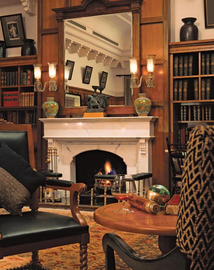 12 BEST Boutique Hotels in Jaipur 46 A cozy library reminiscent of boutique hotels in Jaipur, with bookshelves, a lit fireplace, plush armchairs, a table with brandy and books. A large mirror and ornate vases grace the white mantelpiece.