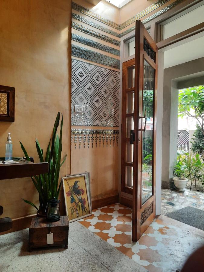 12 BEST Boutique Hotels in Jaipur 17 A wooden door with glass panels opens to a courtyard with plants, evoking the charm of boutique hotels in Jaipur. Inside, a snake plant, framed artwork, a wooden table with a bottle, and patterned tiles adorn the stylish space.